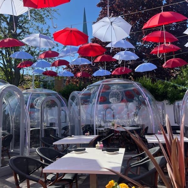 Outdoor dining area with clear igloo-like pods and red, white, and blue umbrellas hanging overhead.
