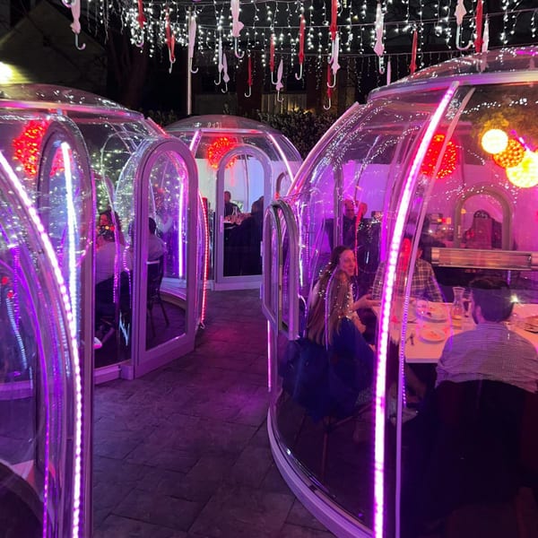 Outdoor dining at night with guests seated inside illuminated clear igloos under string lights and umbrellas.