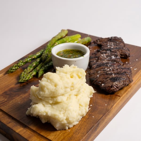 Grilled steak with mashed potatoes, asparagus, and a side of green sauce on a wooden board.