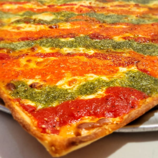 Close-up of a square pizza slice with alternating stripes of red sauce, cheese, and green pesto.