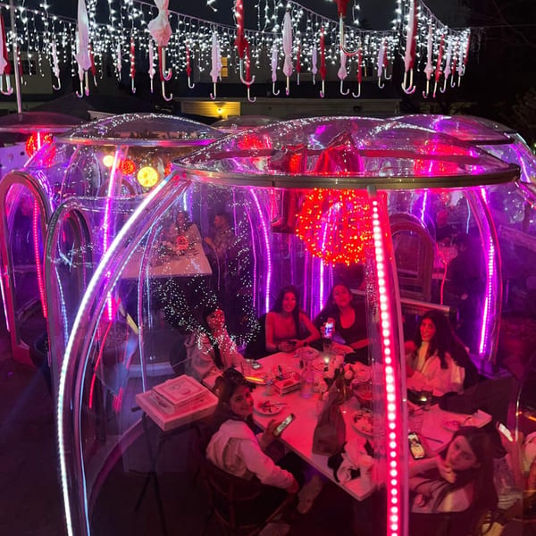 People dining in clear, illuminated outdoor pods under string lights and hanging umbrellas at night.